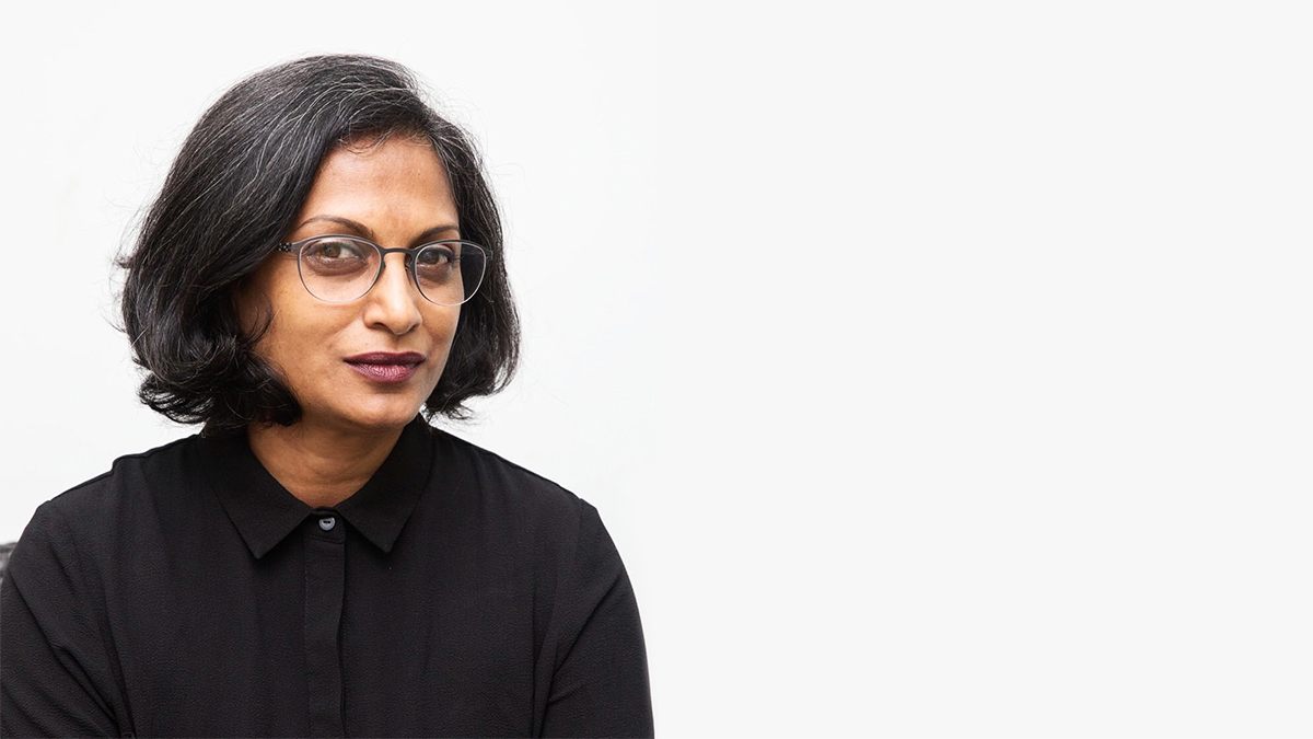 Marina Tabassum, wearing glasses and a black collared shirt, poses seriously against a white backdrop.